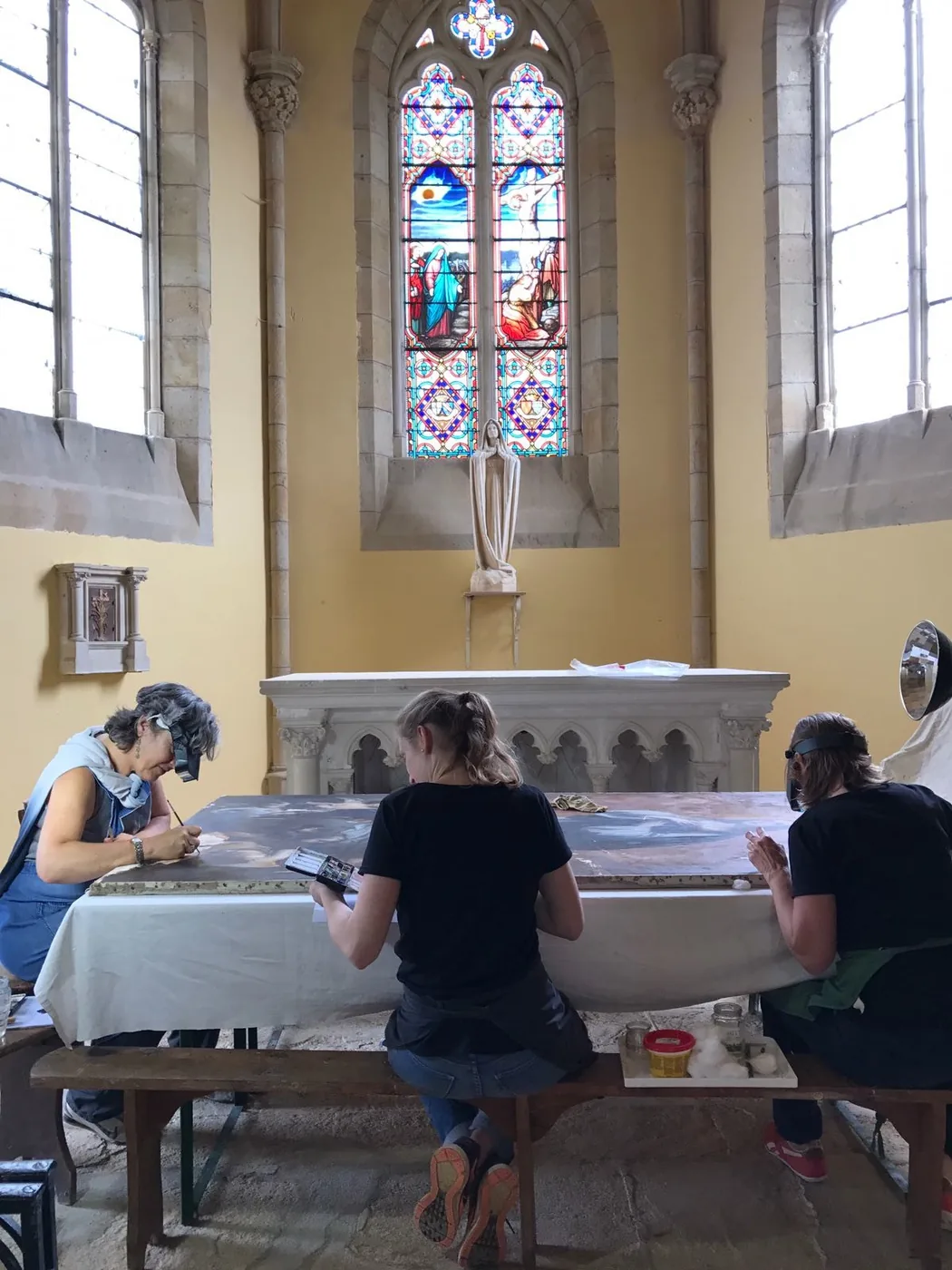 Trois restauratrices travaillent sur un tableau posé à plat, dans une chapelle